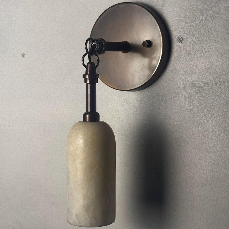 May include: A wall-mounted light fixture with a circular bronze backplate and a dark bronze arm. The fixture features a cream-colored, cylindrical shade suspended from the arm. The fixture is designed for indoor use.