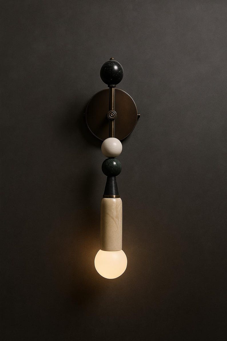 May include: A modern wall sconce with a brass finish and a decorative design. The sconce features a black marble sphere, a white sphere, a green marble sphere, and a cylindrical light fixture with a warm, glowing bulb.