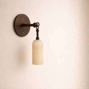 May include: A bronze-toned wall sconce with a circular backplate and a cylindrical, off-white glass shade. The fixture has a simple, elegant design, suitable for various interior styles. The sconce is mounted on a neutral-colored wall.