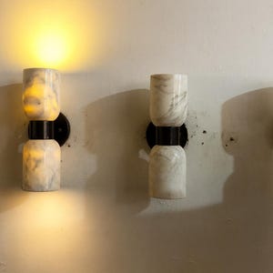 May include: Four wall sconces with a marble-like appearance. Each sconce has two stacked cylindrical sections, connected by a black metal band, and mounted on a black circular base. One sconce is illuminated, casting a warm glow.