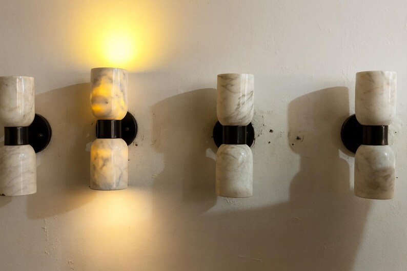 May include: Four wall sconces with a marble-like appearance. Each sconce has two stacked cylindrical sections, connected by a black metal band, and mounted on a black circular base. One sconce is illuminated, casting a warm glow.
