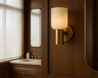 May include: A modern wall sconce with a cylindrical, off-white shade and a gold-colored base. The sconce is mounted on a dark brown wooden wall, with a matching vanity and mirror visible in the background. The light is on.