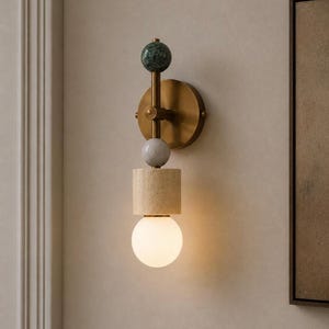 May include: A modern wall sconce with a brass finish, featuring a green marble sphere, a gray marble sphere, and a beige rectangular block. The sconce has a round, warm white light bulb at the bottom. The design is geometric and minimalist.