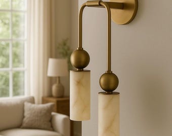 May include: A brass wall sconce with two cylindrical, off-white marble-like shades. The sconce has a circular backplate and curved arms, with a spherical accent above each shade. The fixture is mounted on a light-colored wall.