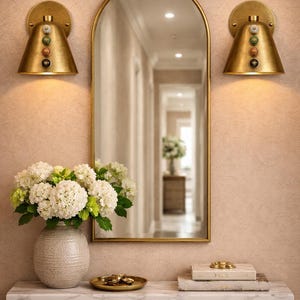 May include: A decorative wall arrangement featuring a gold-framed mirror, two sconces, and a console table. The sconces have a conical shape and are adorned with colorful stones. A vase of white flowers sits on the table.