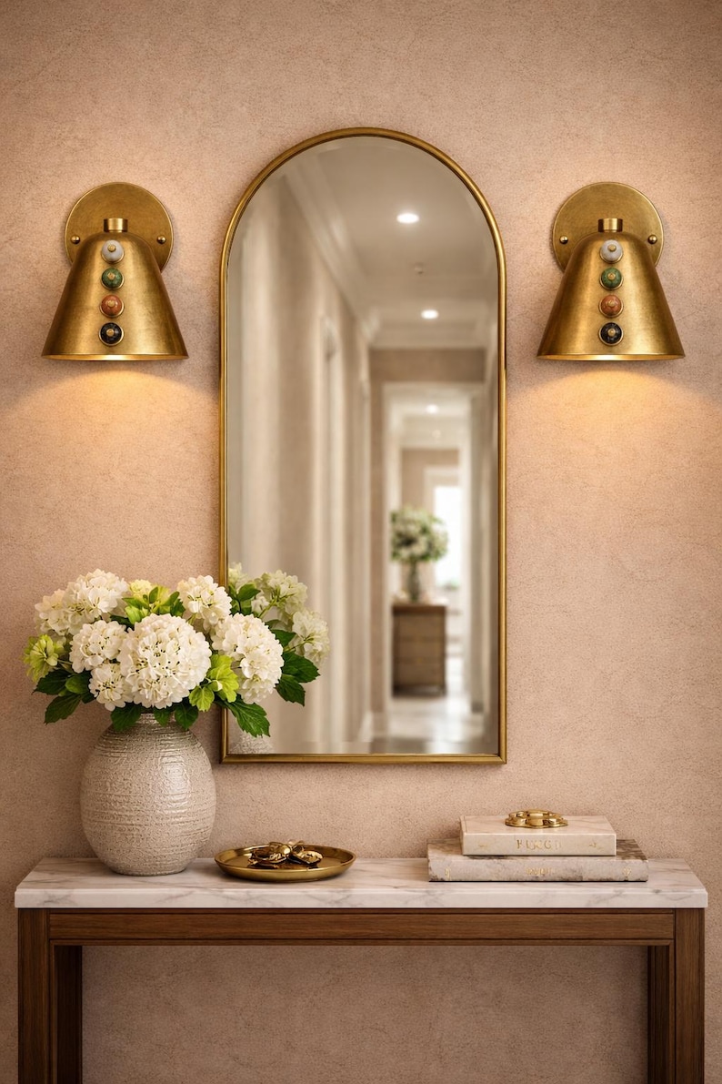 May include: A decorative wall arrangement featuring a gold-framed mirror, two sconces, and a console table. The sconces have a conical shape and are adorned with colorful stones. A vase of white flowers sits on the table.