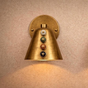 May include: A brass wall sconce with a conical shade and a circular backplate. The sconce features four decorative, colorful, round accents. The light fixture is designed to be mounted on a wall and provides downward illumination.