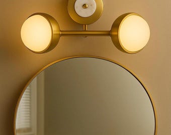 May include: A gold-toned bathroom light fixture with two spherical lights and a central circular mount with a white marble accent. Below, a round gold-framed mirror reflects the room. The fixture is mounted on a beige wall.