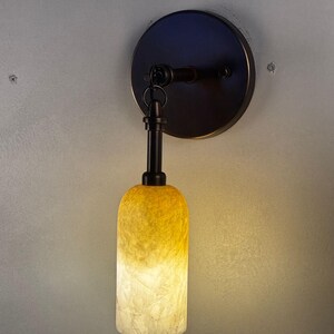 May include: A wall-mounted light fixture with a dark bronze finish. The fixture features a circular base, a curved arm, and a cylindrical shade that transitions from yellow to white. The light is on.