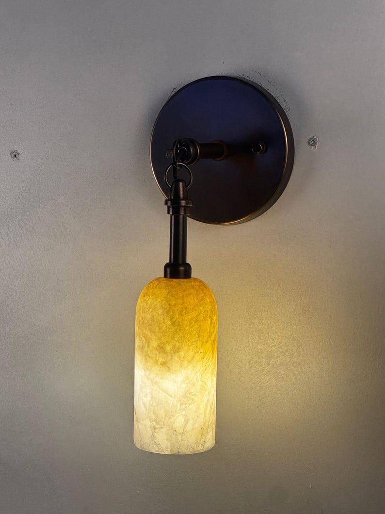 May include: A wall-mounted light fixture with a dark bronze finish. The fixture features a circular base, a curved arm, and a cylindrical shade that transitions from yellow to white. The light is on.