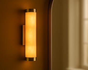 May include: A cylindrical wall sconce with a warm, glowing light. The fixture has a brass finish on the top and bottom, with a cream-colored, translucent center. The sconce is mounted on a matching wall. The overall theme is home decor.