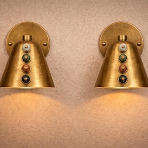 May include: Two brass wall sconces with a conical shade and a circular backplate. Each sconce features a series of colorful decorative accents. The sconces are illuminated, casting a warm glow on the wall. The design is vintage-inspired.