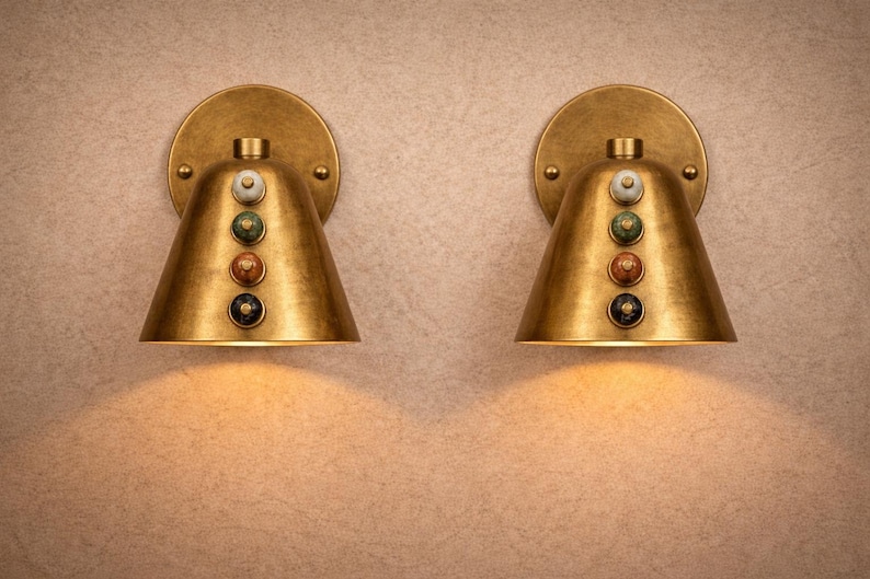 May include: Two brass wall sconces with a conical shade and a circular backplate. Each sconce features a series of colorful decorative accents. The sconces are illuminated, casting a warm glow on the wall. The design is vintage-inspired.