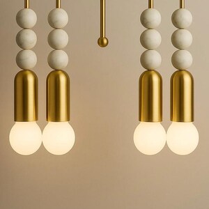 May include: Close-up of modern pendant lights. Each light features a gold-colored cylindrical base, a round, glowing bulb, and a series of white marble spheres. The lights hang from gold-colored rods, creating a stylish and contemporary design.