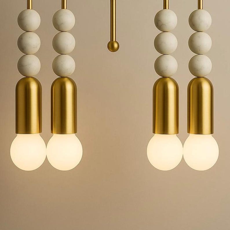May include: Close-up of modern pendant lights. Each light features a gold-colored cylindrical base, a round, glowing bulb, and a series of white marble spheres. The lights hang from gold-colored rods, creating a stylish and contemporary design.
