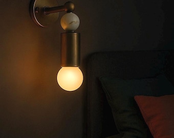 Puede incluir: Una lámpara de pared de latón con base redonda y brazo curvo. Tiene un cuerpo cilíndrico con una esfera de mármol en la parte superior y una bombilla redonda y brillante debajo. La luz ilumina una habitación oscura.