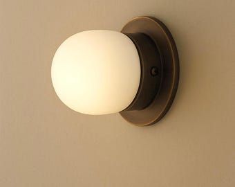 May include: A wall-mounted light fixture featuring a round, frosted white glass globe. The globe is set against a circular, dark bronze-colored metal base. The fixture is designed for indoor use and provides soft, ambient lighting.