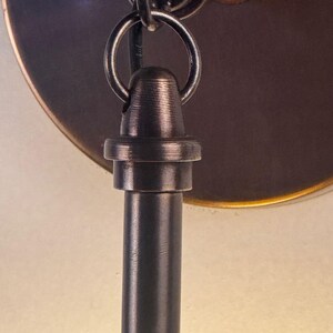 May include: Close-up of a bronze-colored metal light fixture. The fixture features a circular mounting plate, a decorative ring, and a cylindrical rod. The metal has a textured finish, and a small, rusty-looking screw is visible.