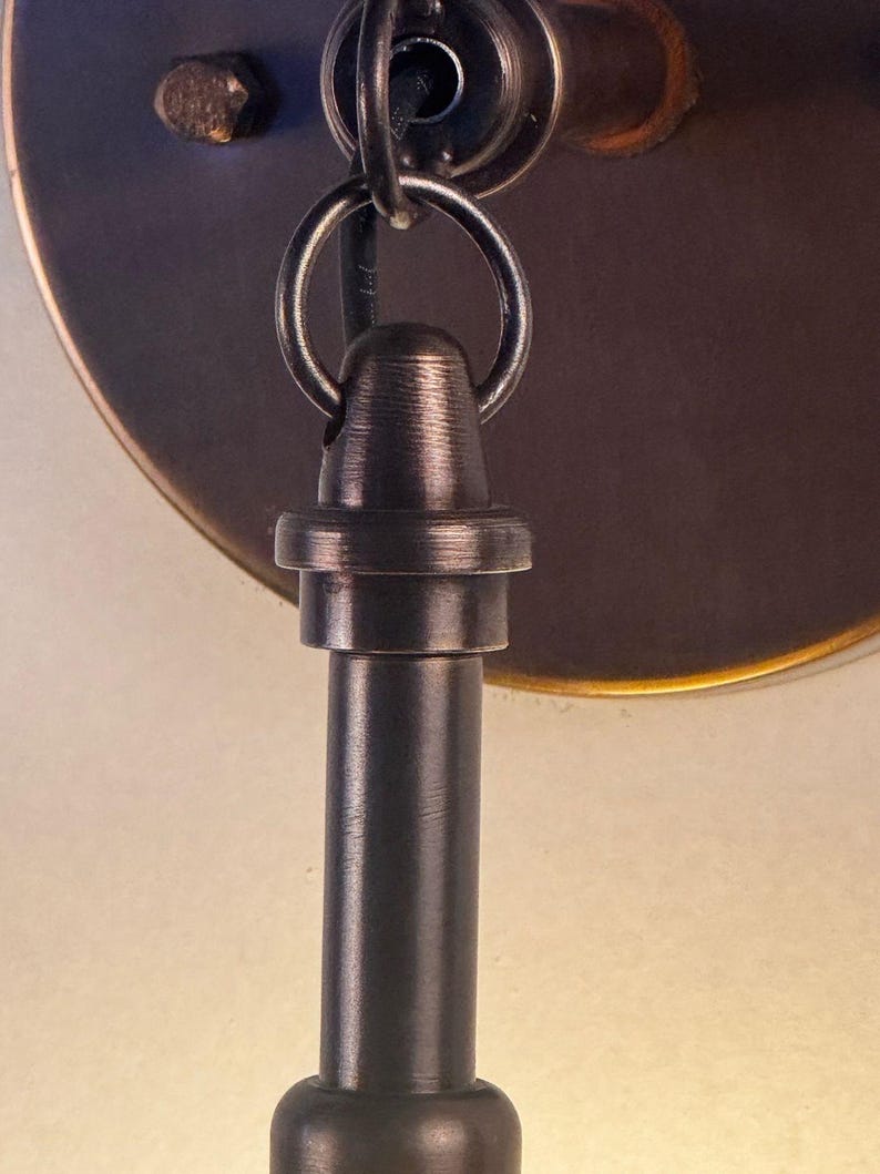 May include: Close-up of a bronze-colored metal light fixture. The fixture features a circular mounting plate, a decorative ring, and a cylindrical rod. The metal has a textured finish, and a small, rusty-looking screw is visible.
