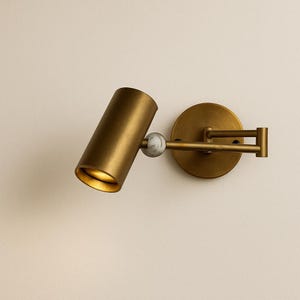 May include: A brass-toned wall-mounted spotlight fixture. The cylindrical light head is connected to a round base with an adjustable arm. A small, marble-like sphere is between the light and the arm. The light is angled downward, illuminating the interior.