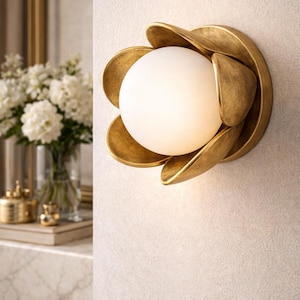 May include: A gold-colored wall sconce with a floral design. The sconce features a central white globe light surrounded by petal-shaped elements. The fixture is mounted on a light-colored wall, creating a warm, inviting ambiance.