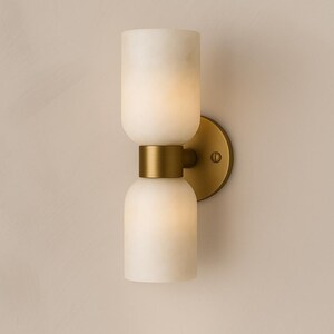 May include: A modern wall sconce with a double-layered, off-white, cylindrical shade. The shade is connected by a brass-colored band and mounted on a round brass-colored backplate. The sconce is designed for indoor lighting.