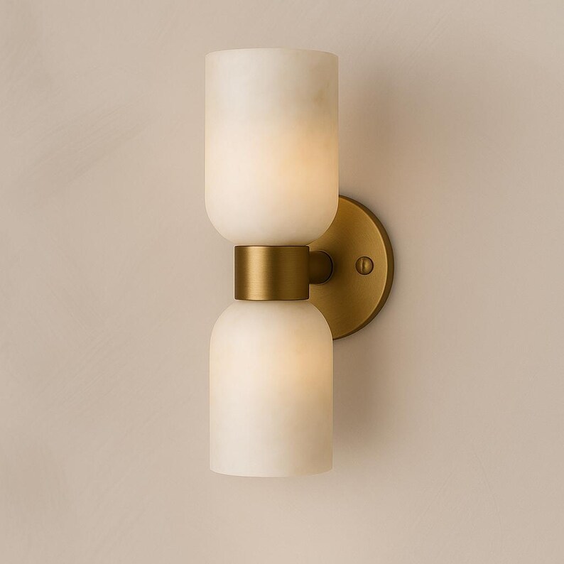 May include: A modern wall sconce with a double-layered, off-white, cylindrical shade. The shade is connected by a brass-colored band and mounted on a round brass-colored backplate. The sconce is designed for indoor lighting.