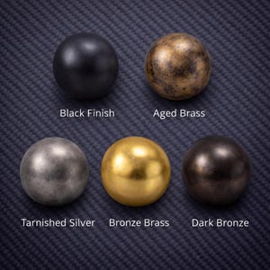 May include: Five round cabinet knobs in various finishes: black, aged brass, tarnished silver, bronze brass, and dark bronze. Each knob is displayed on a textured, dark blue surface, with the finish name below each knob.
