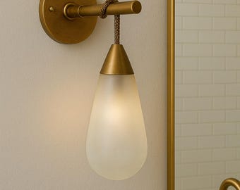 May include: A wall-mounted light fixture featuring a circular brass backplate, a horizontal arm, and a teardrop-shaped frosted glass shade suspended by a textured cord. The metal components have a brushed brass finish, and the light emits a soft glow.
