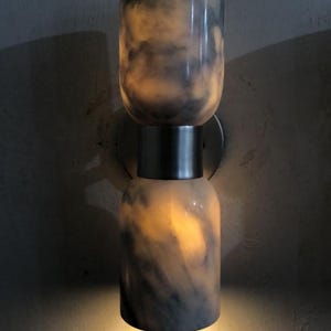 May include: A modern wall sconce with a cylindrical design. The fixture is composed of two stacked, marble-like cylinders with a dark gray and white pattern. The sconce emits a warm, inviting light.