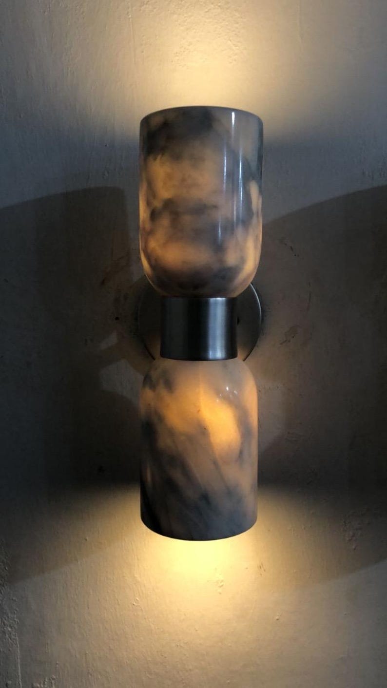 May include: A modern wall sconce with a cylindrical design. The fixture is composed of two stacked, marble-like cylinders with a dark gray and white pattern. The sconce emits a warm, inviting light.