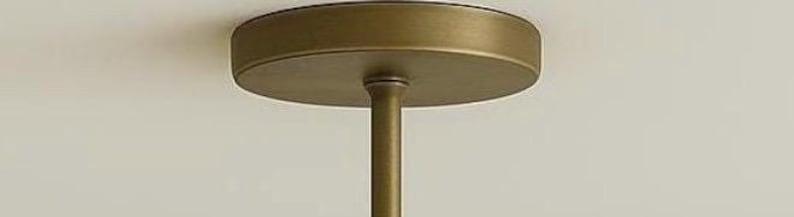 May include: A close-up view of a brass-colored ceiling light fixture. The fixture has a circular base and a central rod extending downwards. The light fixture is mounted on a white ceiling.