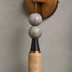 May include: A modern wall sconce with a brass finish, featuring a series of spherical and cylindrical elements. The design includes a marble-like sphere at the top, two gray spheres, and a light-colored cylindrical base with a white globe at the bottom.