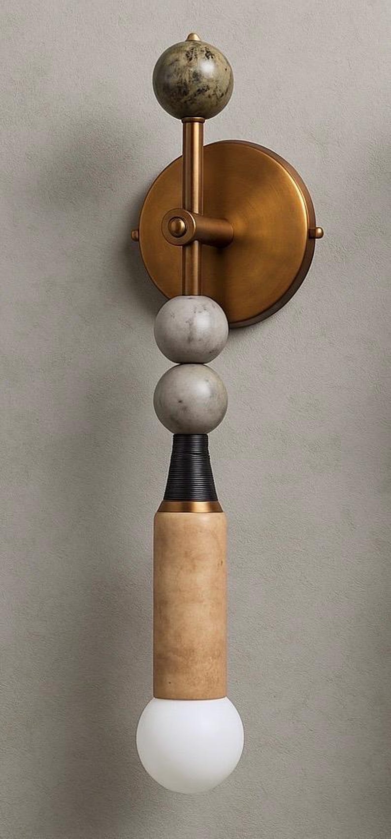 May include: A modern wall sconce with a brass finish, featuring a series of spherical and cylindrical elements. The design includes a marble-like sphere at the top, two gray spheres, and a light-colored cylindrical base with a white globe at the bottom.
