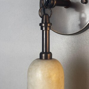 May include: A wall-mounted light fixture featuring a cylindrical, translucent, off-white stone shade with a textured surface. The shade is suspended from a dark bronze-toned metal arm and circular wall plate by a small metal ring.