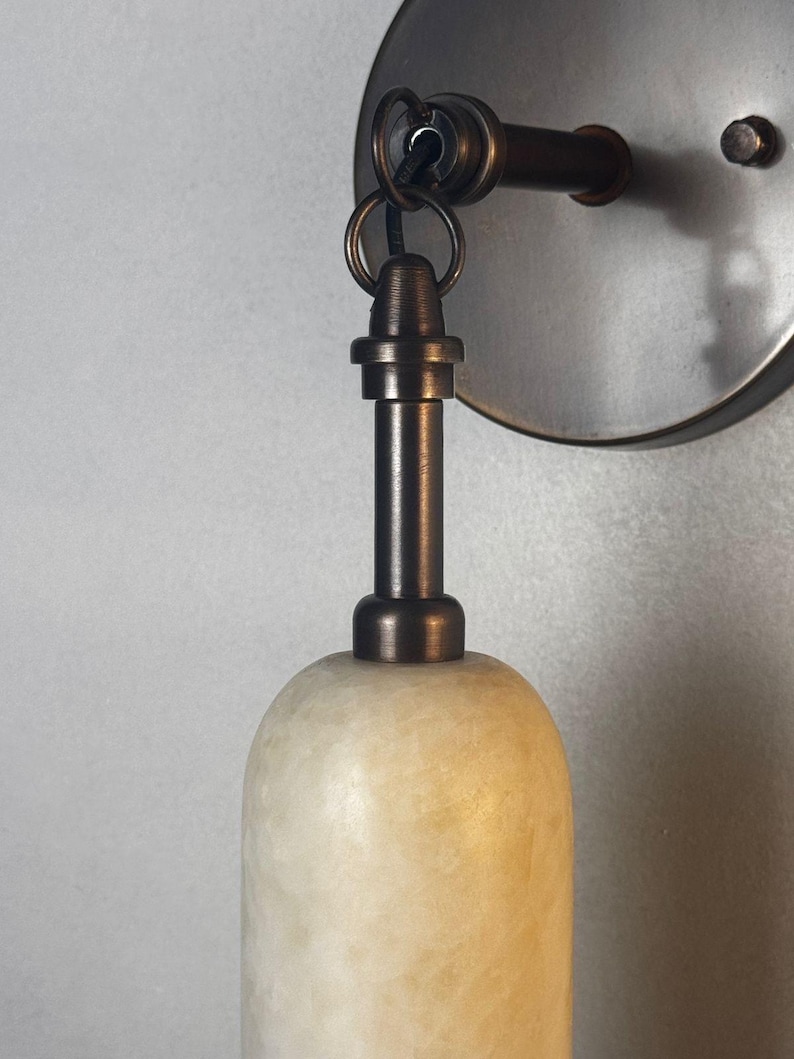 May include: A wall-mounted light fixture featuring a cylindrical, translucent, off-white stone shade with a textured surface. The shade is suspended from a dark bronze-toned metal arm and circular wall plate by a small metal ring.