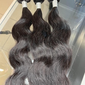 Puede incluir: Tres paquetes de extensiones de cabello largo, ondulado y de color marrón oscuro. Cada paquete está asegurado en la parte superior con un grueso hilo de color claro, que da paso a ondas sueltas y fluidas que caen con una textura uniforme.
