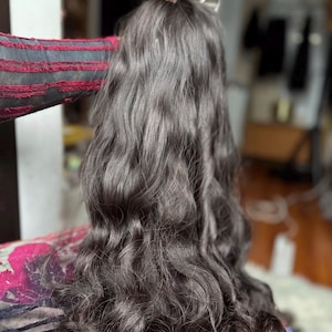 Könnte beinhalten: Eine lange, dunkelbraune Perücke mit lockeren, fließenden Wellen, die den Rücken hinunterfallen. Das Haar hat ein natürliches, mehrfarbiges Aussehen mit subtilen Highlights, die Tiefe und Textur über die gesamte Länge der Wellen erzeugen.