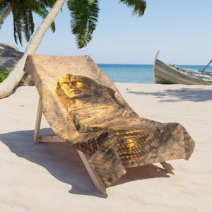 Peut inclure: Chaise de plage en bois avec une assise en tissu représentant un Sphinx. Le Sphinx a des yeux dorés et se détache sur une scène de plage ensoleillée et sablonneuse. Un palmier et un bateau sont visibles en arrière-plan.