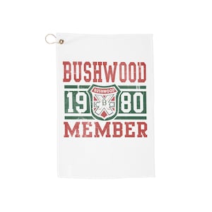 Puede incluir: Toalla de golf blanca con el texto "BUSHWOOD 1980 MEMBER" en rojo y verde. La toalla tiene un ojal metálico en la esquina superior izquierda. El diseño incluye un escudo con las letras "CBC" y una cruz.