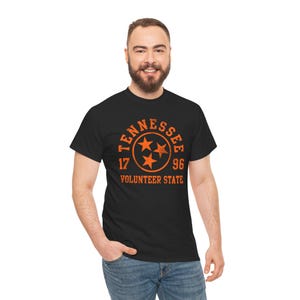 May include: Black t-shirt with an orange graphic. The graphic includes the word "Tennessee" in a circular design, three stars, the numbers "17" and "96", and the words "Volunteer State". The shirt is made of a soft material.