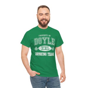 Puede incluir: Una camiseta verde con el texto blanco "PROPERTY OF DOYLE XXL DRINKING TEAM". El diseño incluye tréboles y una fuente de estilo universitario. La camiseta tiene cuello redondo y mangas cortas.
