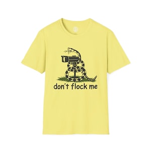 Puede incluir: Una camiseta de manga corta de color amarillo pálido con un gráfico en blanco y negro de una serpiente enroscada alrededor de una cámara de seguridad sobre un parche de hierba verde. Debajo, el texto "don't flock me" en fuente negra redondeada.