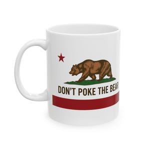 May include: White ceramic mug featuring the California state flag design. The mug displays a brown bear, a red star, and a red stripe, with the text "DON'T POKE THE BEAR" in black. The mug has a white handle.
