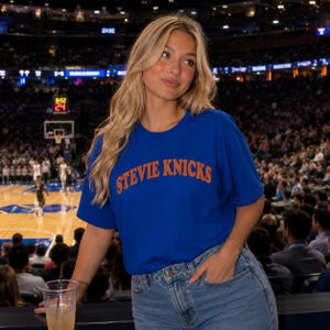 Peut inclure: Un t-shirt bleu roi à manches courtes avec le texte STEVIE KNICKS imprimé en orange dans une police arquée sur la poitrine, associé à un jean bleu clair. Le t-shirt présente une encolure ronde et une coupe standard, porté dans un stade.