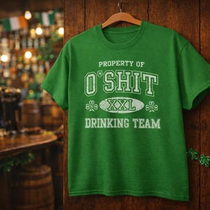 Puede incluir: Una camiseta verde con el texto "PROPERTY OF O'SHIT XXL DRINKING TEAM" en letras blancas. La camiseta está colgada en una percha de madera sobre un fondo de madera. El fondo incluye un bar.