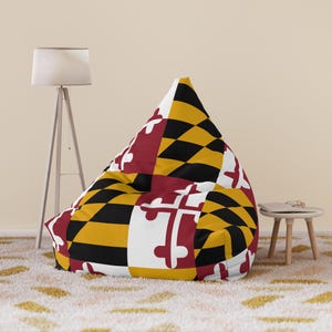 Może przedstawiać: Duży worek do siedzenia z wzorem flagi stanu Maryland. Worek ma trójkątny kształt i pokryty jest wzorem w kolorze czerwonym, białym, czarnym i złotym. Krzesło stoi na wzorzystym dywanie.