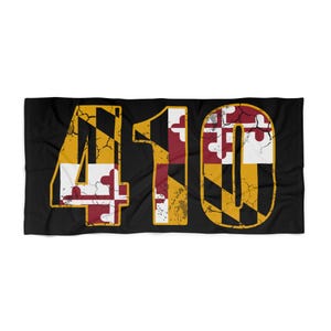 May include: Black rectangular towel featuring the number "410" in a distressed, textured design. The numbers are filled with the Maryland state flag's colors: gold, white, red, and black. The towel is likely for home or beach use.