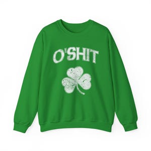 Puede incluir: Sudadera verde con la frase "O'SHIT" en blanco, sobre un trébol blanco desgastado. La sudadera es de color verde liso, con cuello redondo y mangas largas. El diseño tiene un aspecto vintage.