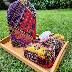 Pode incluir: Um conjunto decorativo de chaleira e caixa de lenços. O conjunto apresenta um design de patchwork com cores vibrantes, incluindo vermelho, roxo, amarelo e preto. A chaleira é alta e arredondada, enquanto a caixa de lenços é retangular. Ambos estão em uma bandeja de madeira.